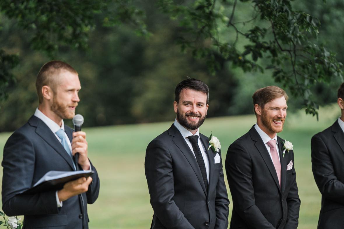 Tom Allen officiating an outdoor garden wedding ceremony as the groom and groomsmen smile