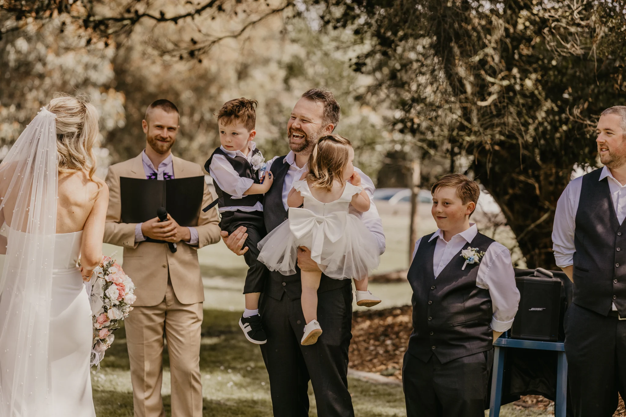 Tom Allen officiating an outdoor garden wedding ceremony as the groom and groomsmen smile