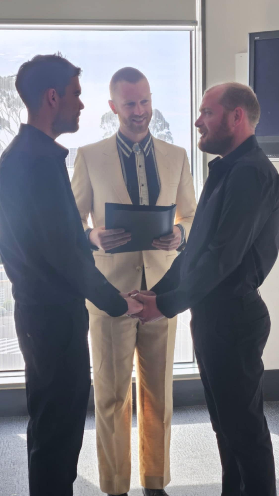 Tom Allen officiating a same-sex wedding ceremony, two grooms holding hands at an intimate indoor venue