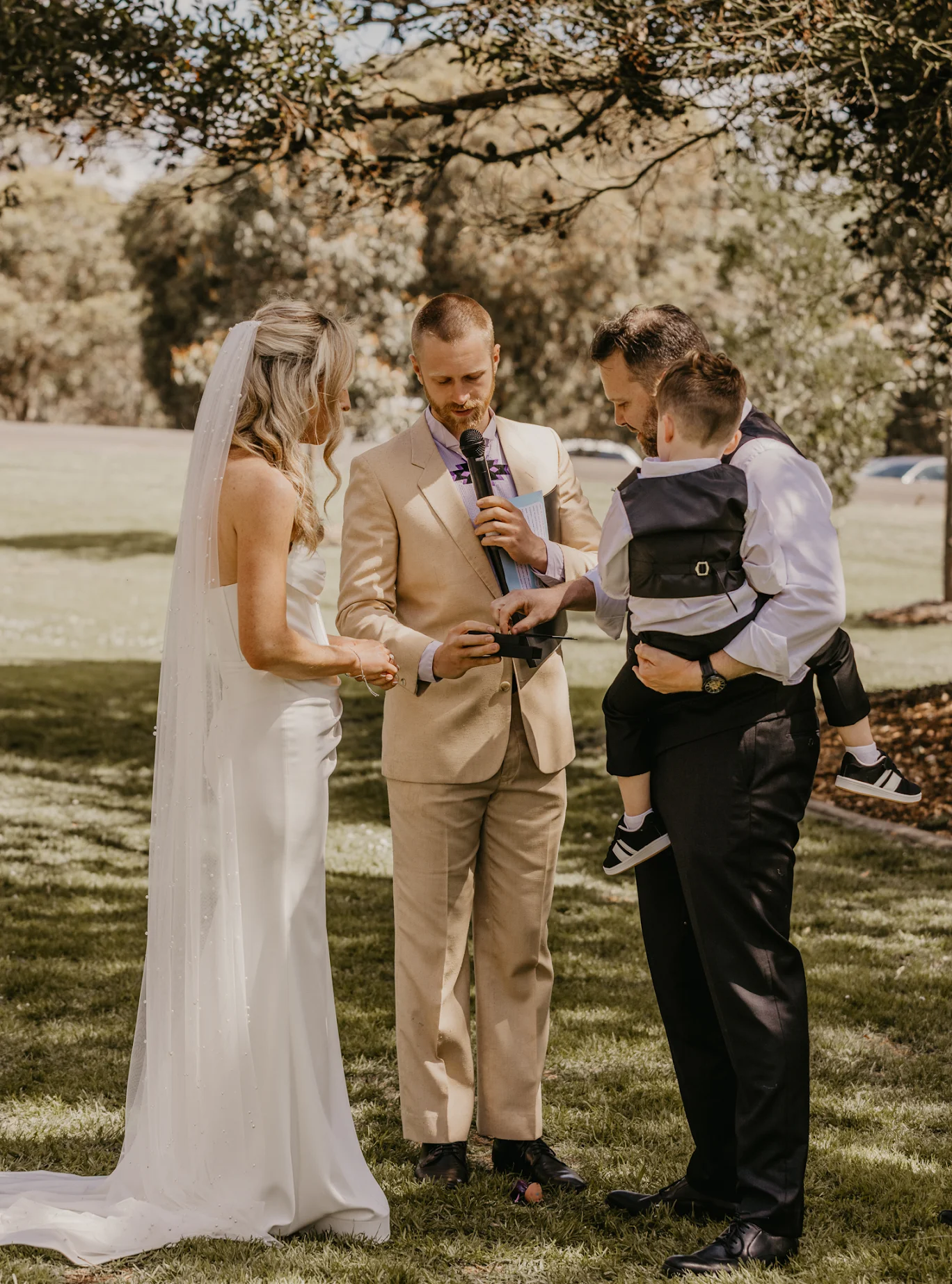 Tom Allen officiating an outdoor garden wedding, reading from his folder as a groom holds a young child and the bride stands opposite