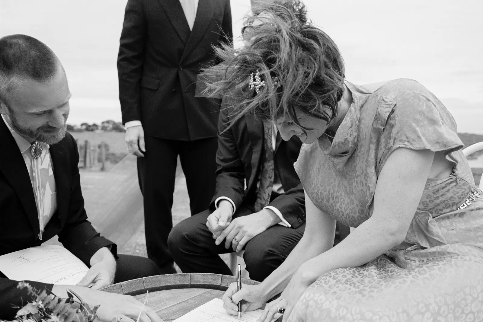 Black and white photo of a bride signing the marriage certificate on a wine barrel outdoors on a windy day, with Tom Allen watching beside her