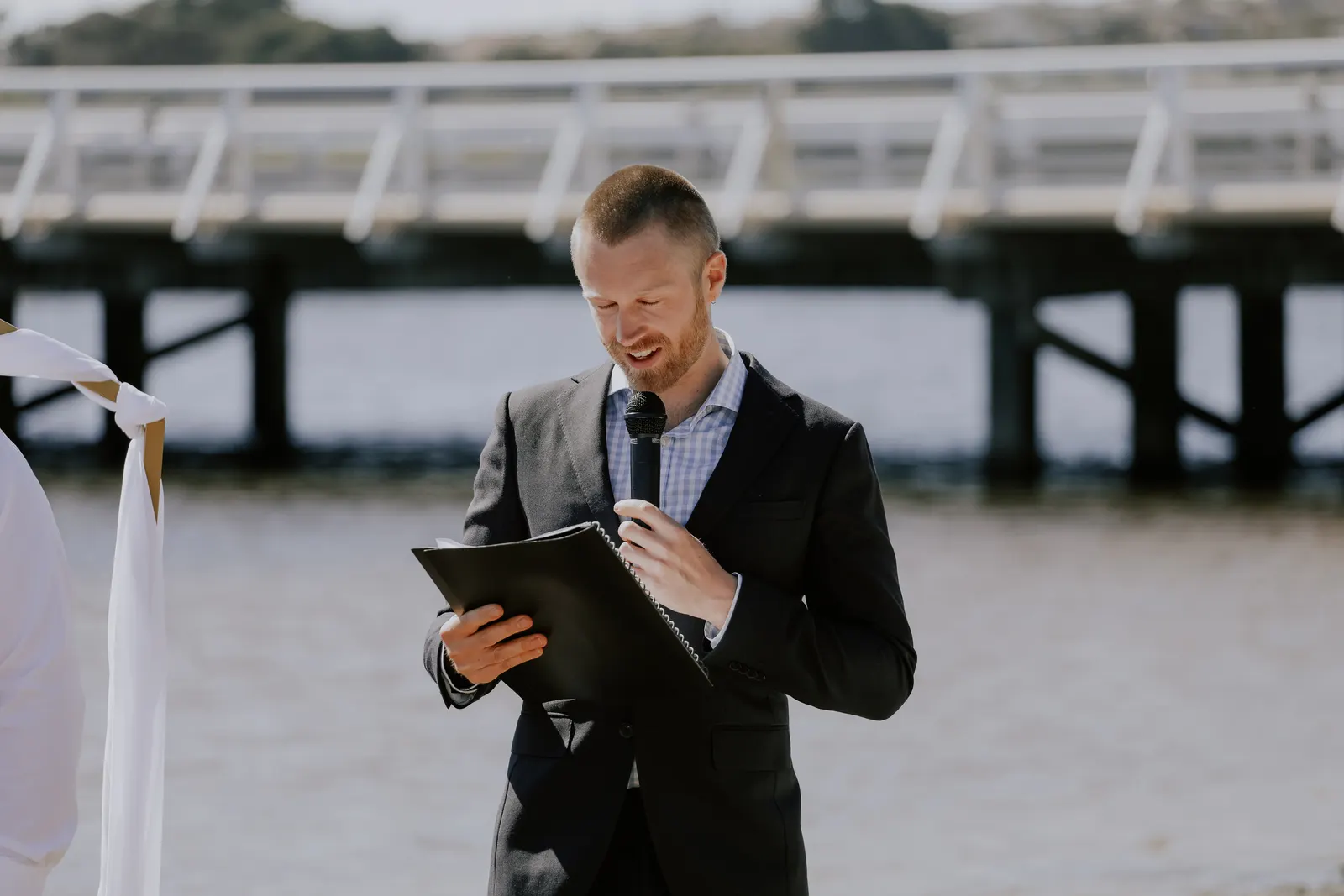 Tom Allen reading from his ceremony folder with a smile at a waterfront ceremony, a long pier stretching across the water behind him