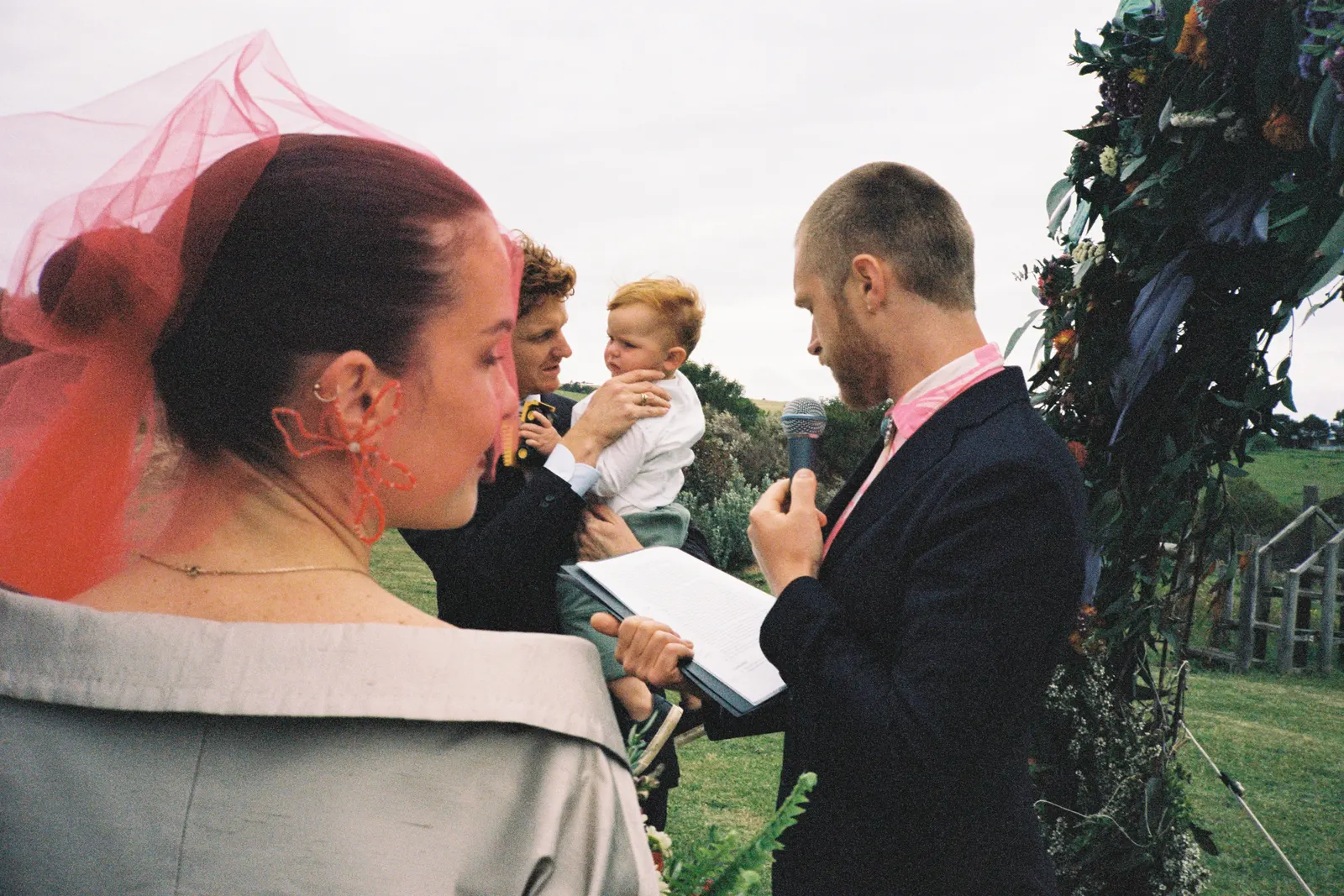 Wide shot of a wedding ceremony in a sunlit bush clearing, guests seated in a circle around the couple among tall eucalyptus trees