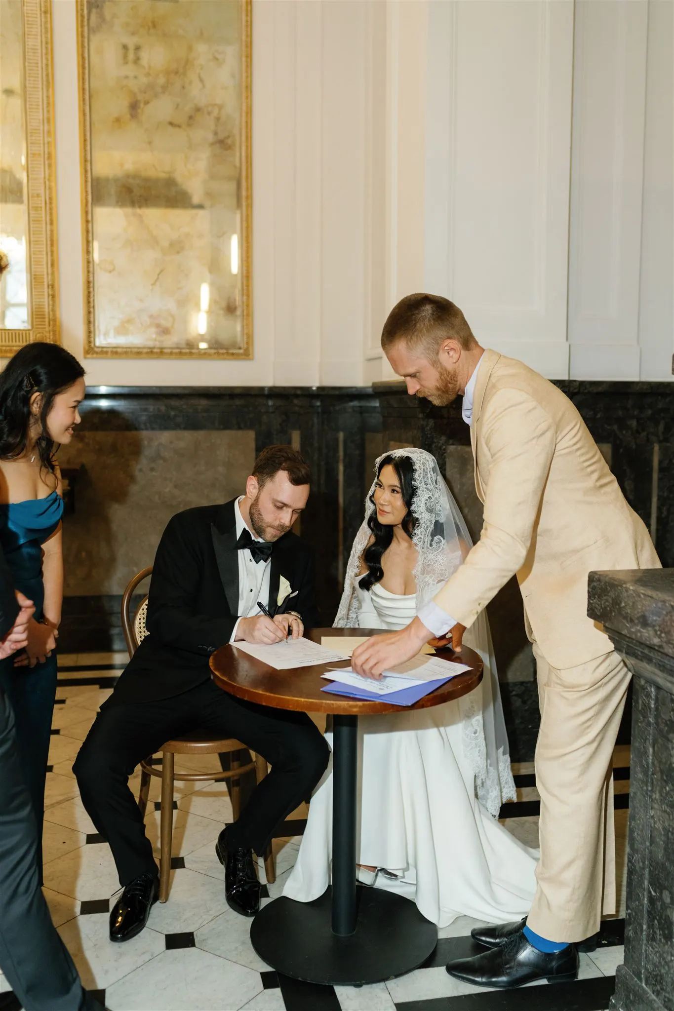 A bride and groom signing the marriage register at a small round table inside an elegant venue with marble floors and a gold-framed mirror, watched by two witnesses