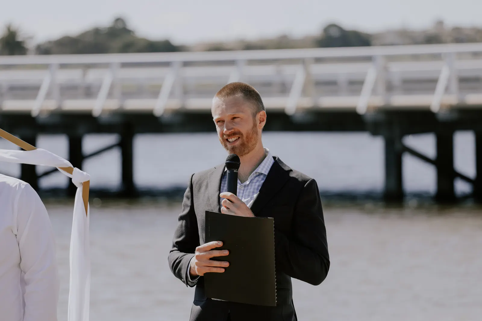 Tom Allen smiling at the couple during a waterfront ceremony, holding his ceremony folder with a jetty stretching across the water behind him