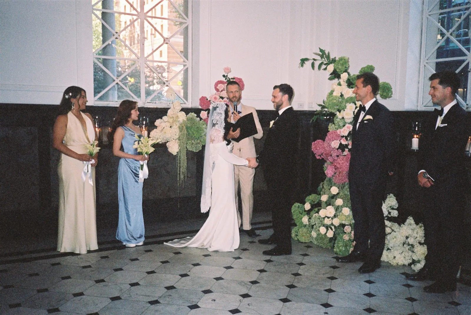 A bride and groom exchanging vows in a sunlit garden clearing, officiated by Tom Allen with their wedding party gathered around them