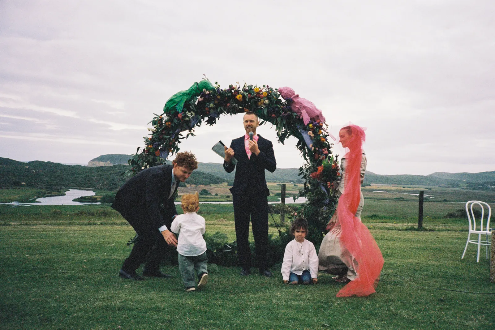 Tom Allen officiating a ceremony under a colourful floral arch on a hilltop farm, with a river valley and rolling hills stretching into the distance behind the couple and their children