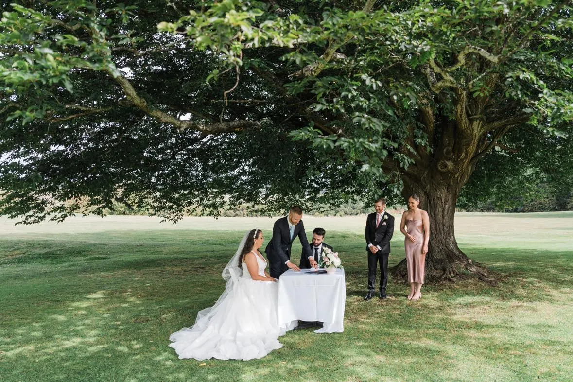 A couple signing the marriage register at a table beneath a large tree, with Tom Allen and their witnesses looking on