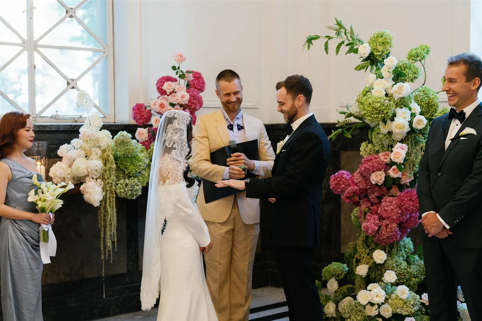 A groom laughing as he holds two young children at the altar during the ceremony, with Tom Allen reading in the background and the bride facing the group