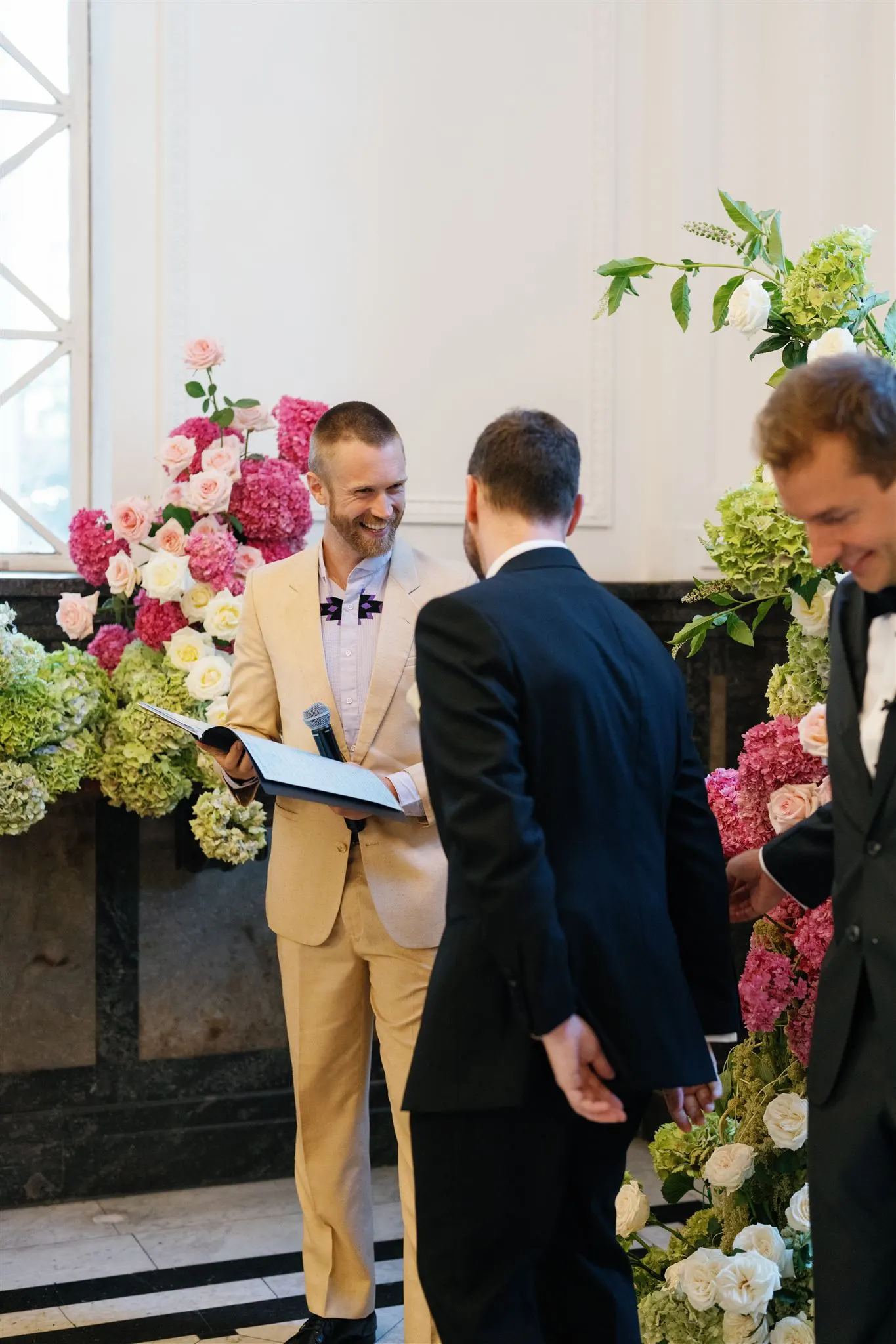 Close-up of Tom Allen reading the ceremony to a couple at a colourful floral arch as the groom holds a baby