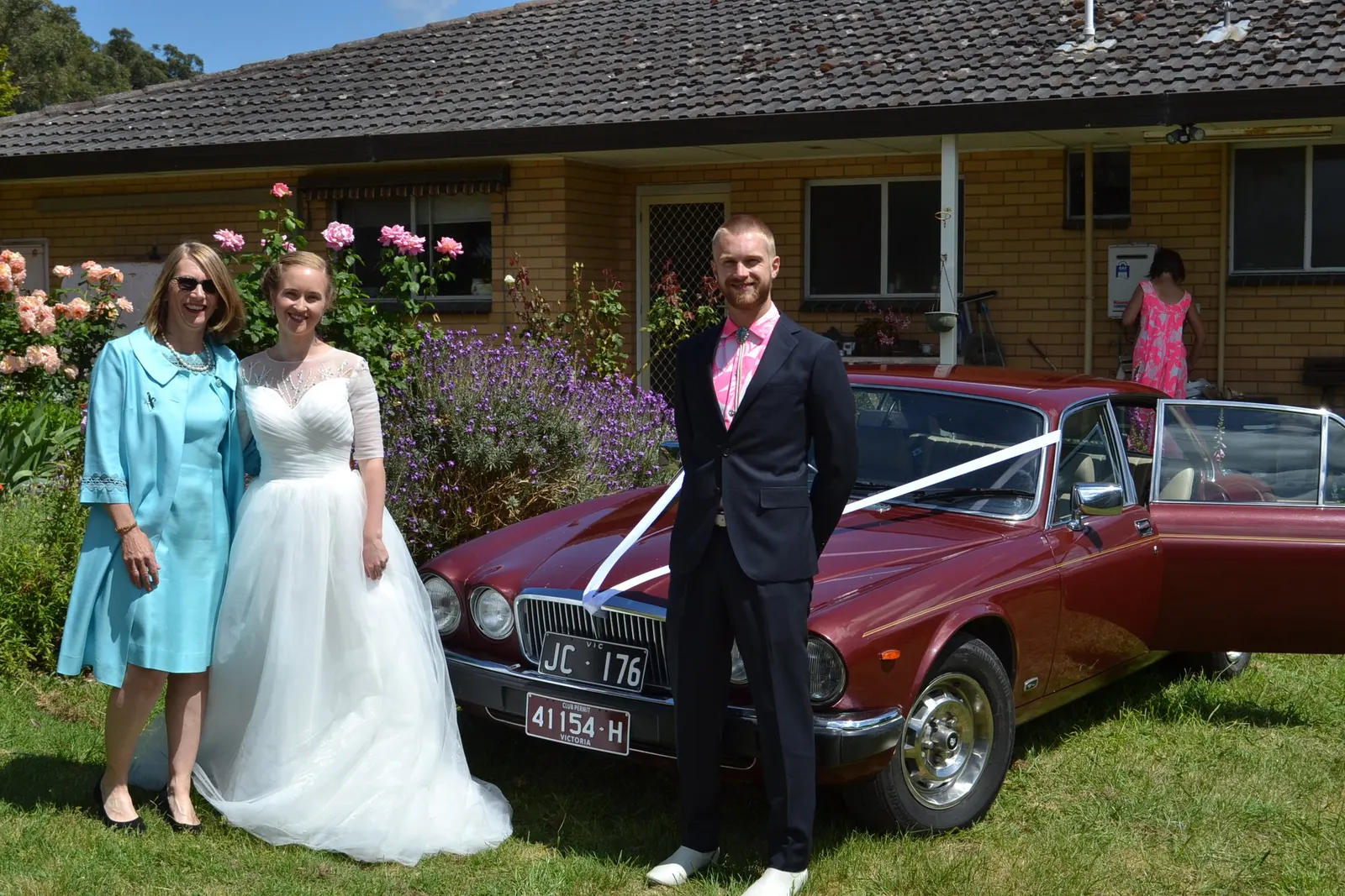 Tom Allen with a bride, groom, and family member beside a vintage red car decorated with white ribbons
